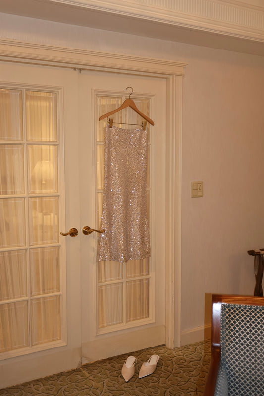 A sparkling champagne gold sequined skirt hanging on a wooden hanger in a doorway, with a pair of white pointed kitten heels on the carpet below in an indoor room setting.