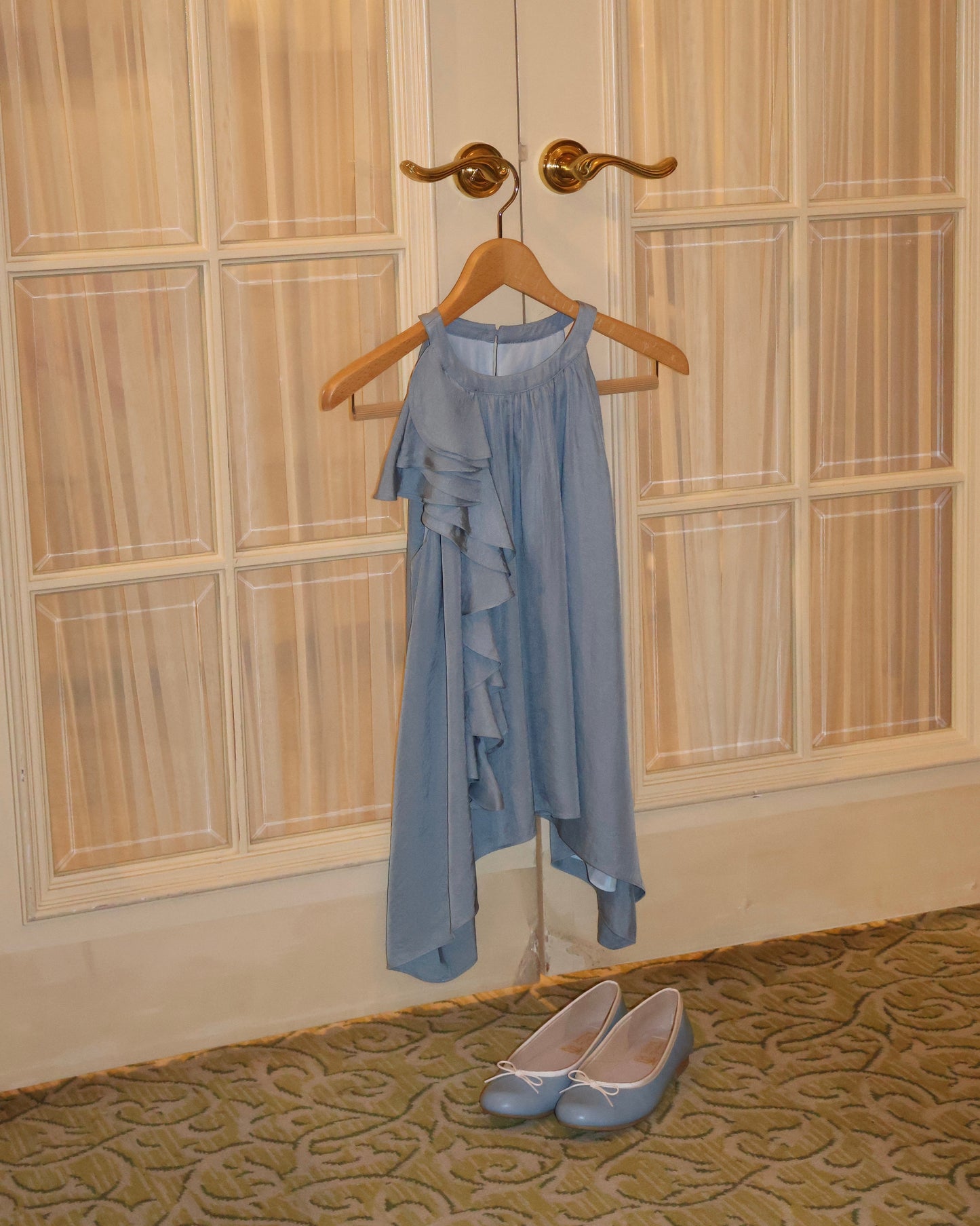 A light dusty blue sleeveless top with ruffle details hangs on a wooden hanger from a brass door hook in front of cream paneled doors, with a pair of matching light blue ballet flats on the patterned carpet below.