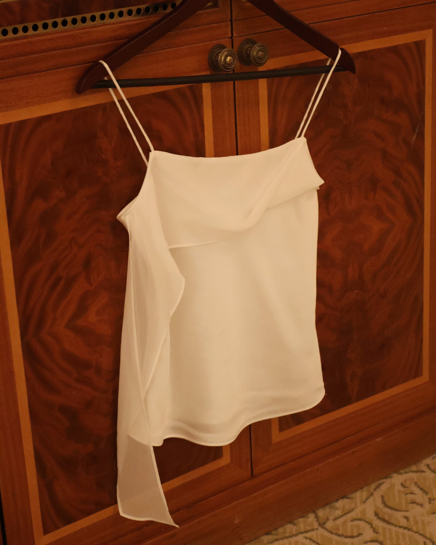 A white camisole top with thin straps hanging on a dark wooden hanger against a polished wood cabinet door.