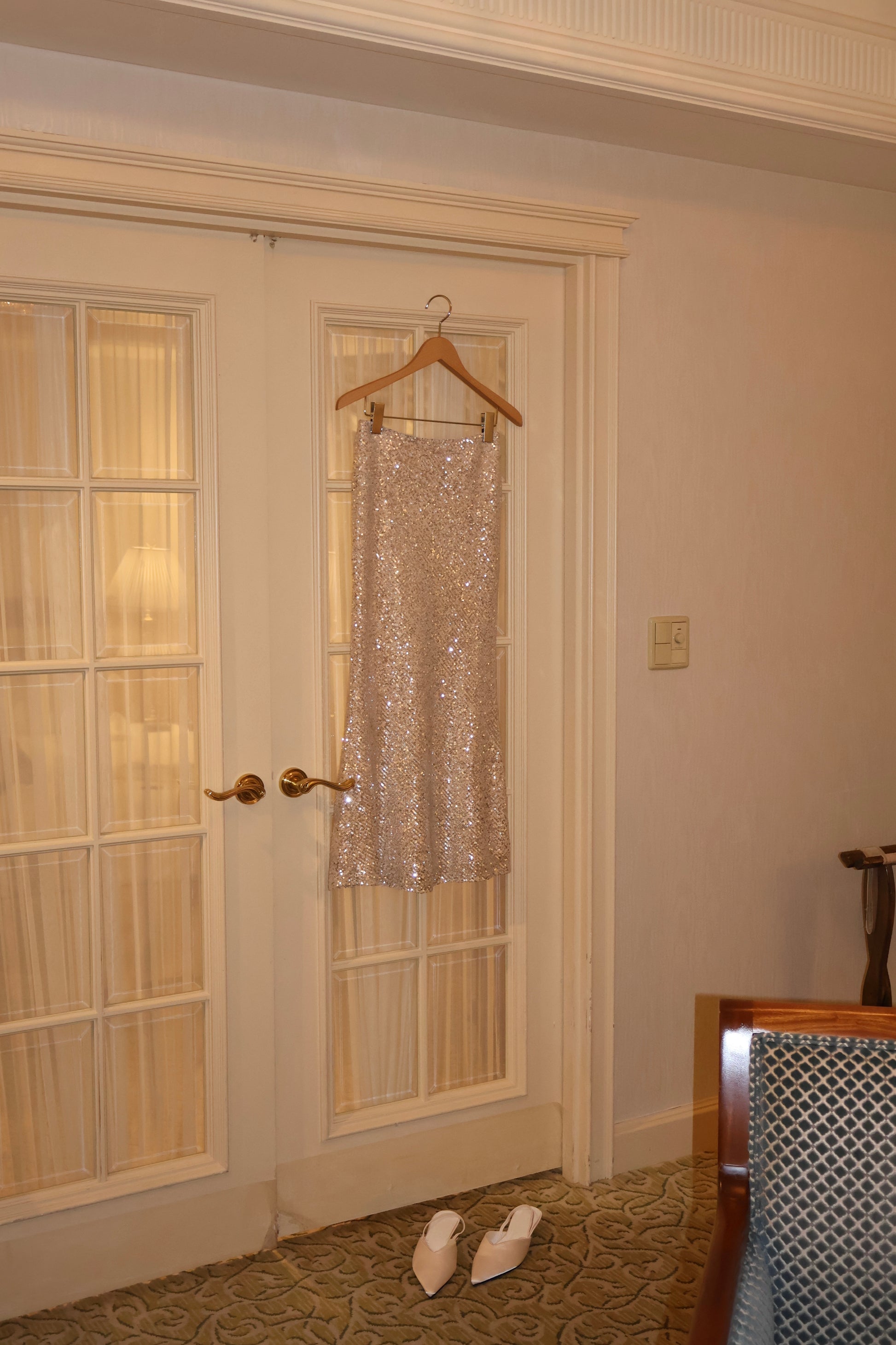 A sparkling champagne gold sequined skirt hanging on a wooden hanger in a doorway, with a pair of white pointed kitten heels on the carpet below in an indoor room setting.