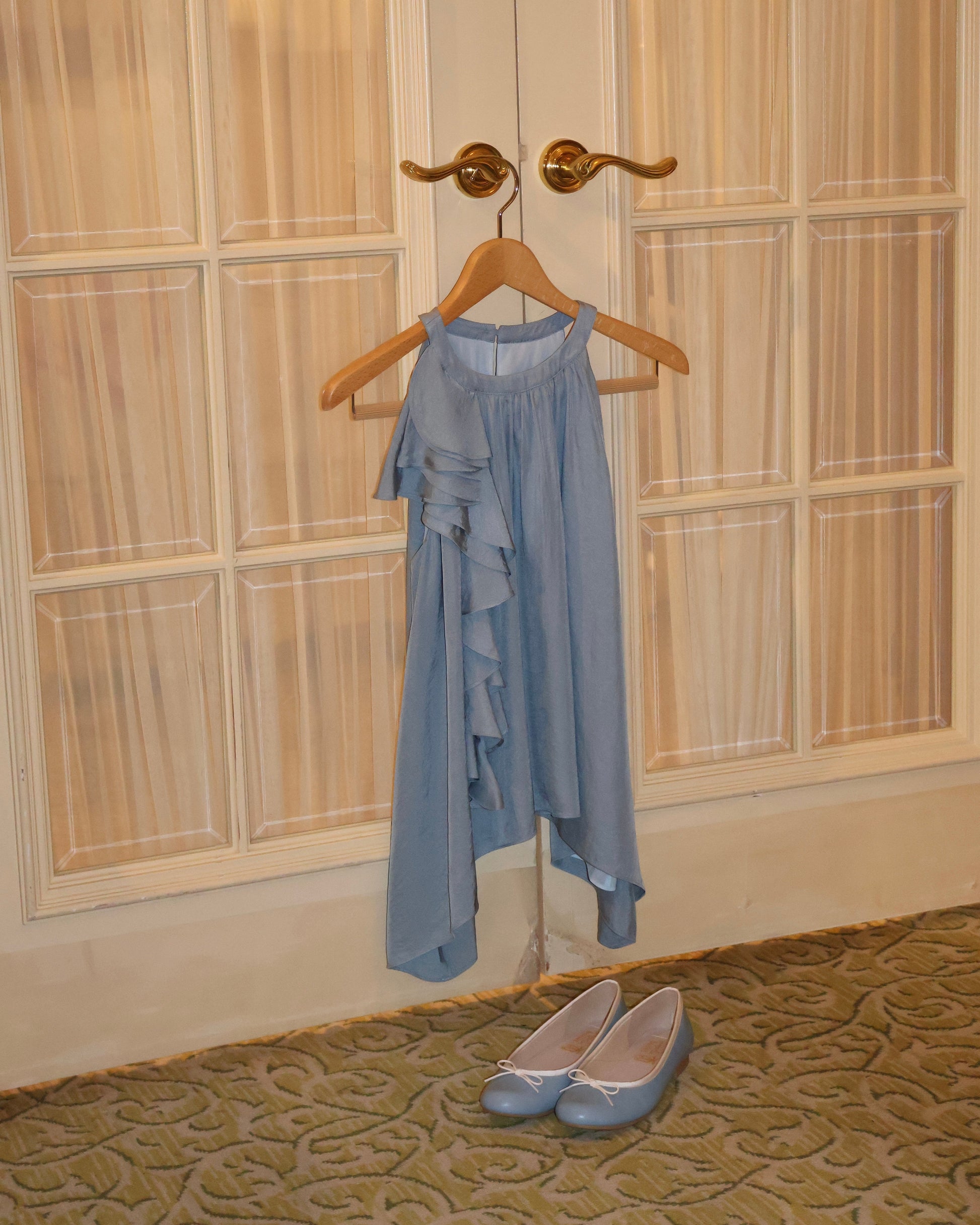 A light dusty blue sleeveless top with ruffle details hangs on a wooden hanger from a brass door hook in front of cream paneled doors, with a pair of matching light blue ballet flats on the patterned carpet below.