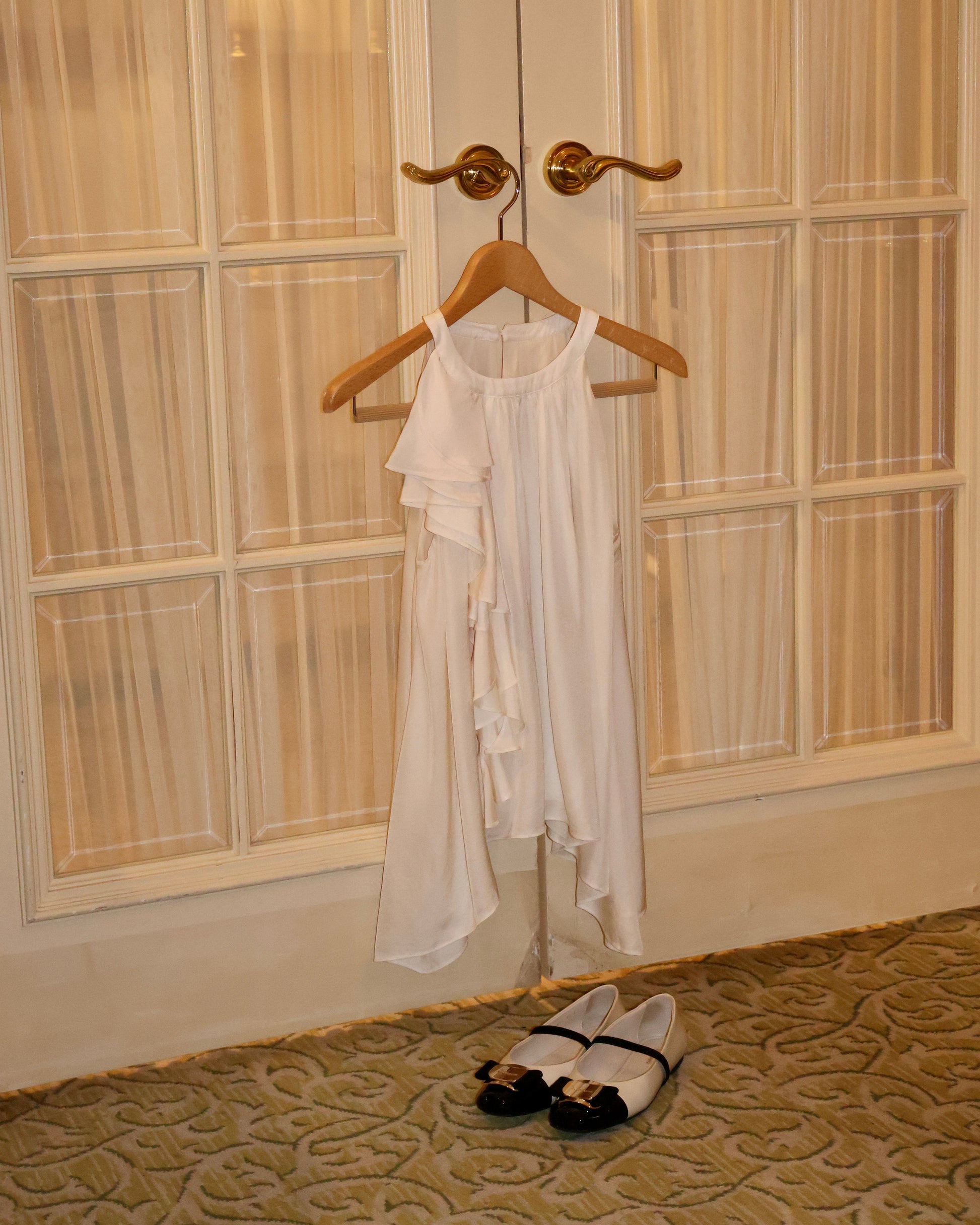 A creamy sleeveless ruffled top hangs on a wooden hanger from a brass door hook in front of cream paneled doors, with a pair of black and white ballet flats placed on the patterned carpet below.
