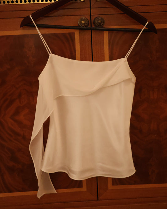 A white satin camisole top with thin spaghetti straps and an asymmetrical draped hem, hanging on a dark wooden hanger against a warm-toned wooden door background.