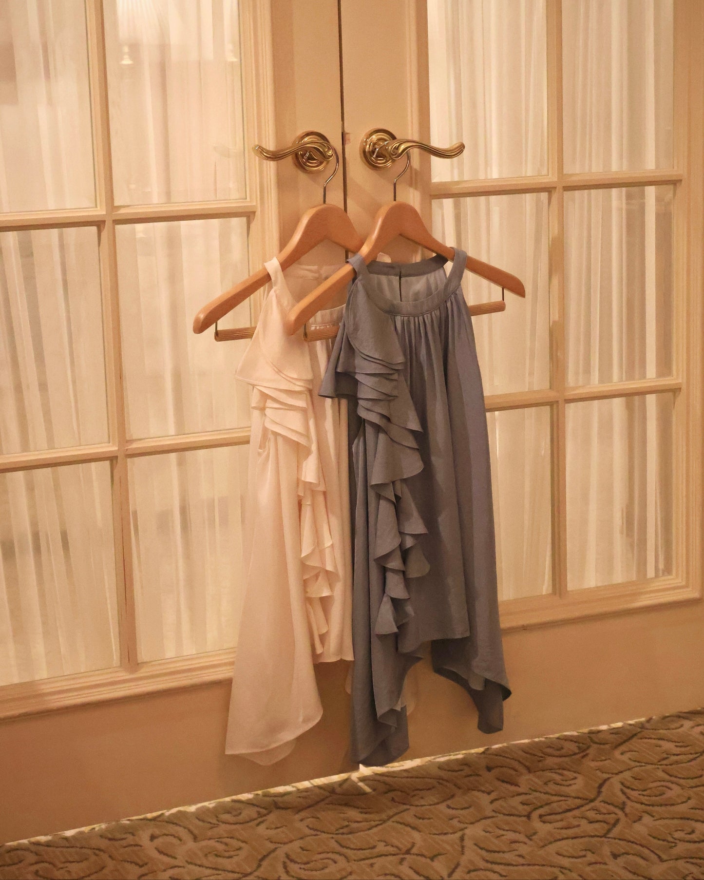 Two sleeveless ruffled tops hanging on wooden hangers against a set of double doors with sheer curtains. One top is a very light vanilla yellow and the other is a slate blue.