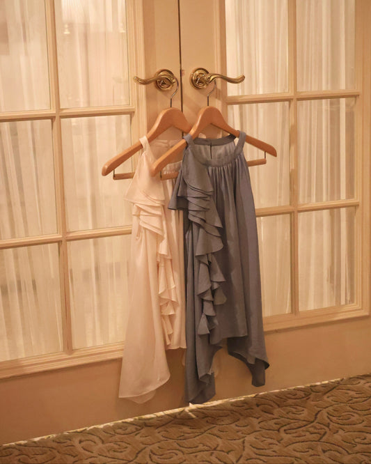 Two sleeveless ruffled tops hanging on wooden hangers against a set of double doors with sheer curtains. One top is a very light vanilla yellow and the other is a slate blue.