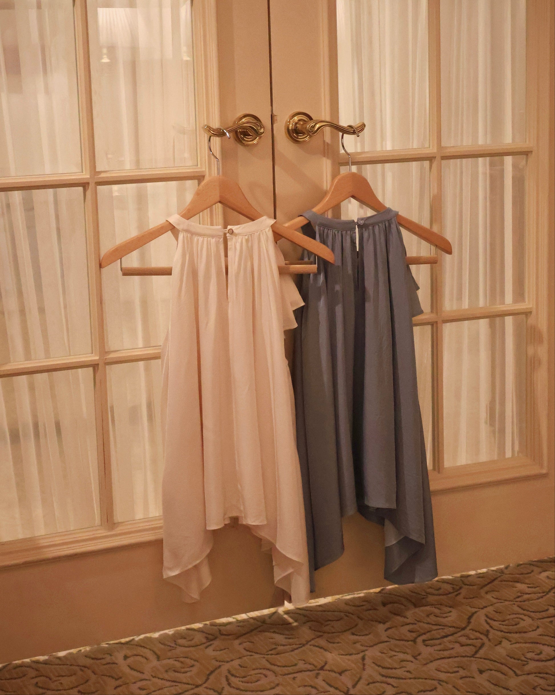 Two sleeveless draped tops hanging on wooden hangers on a double door in a warm-toned room. The top on the left is a very light vanilla yellow, and the top on the right is a dusty slate blue.