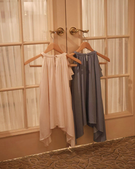 Two sleeveless draped tops hanging on wooden hangers on a double door in a warm-toned room. The top on the left is a very light vanilla yellow, and the top on the right is a dusty slate blue.